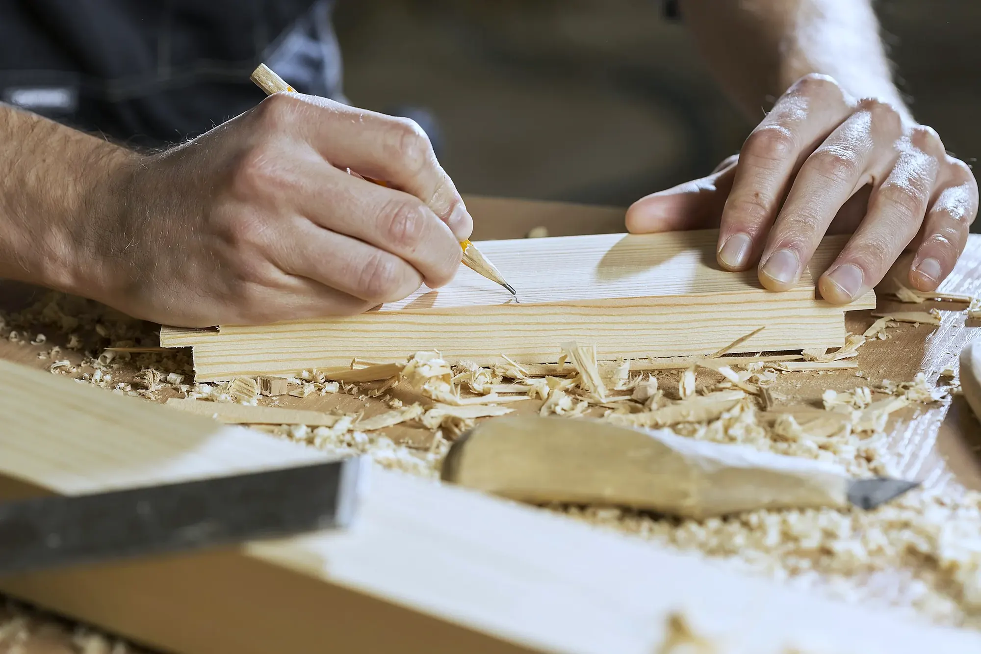 Main traçant un repère au crayon sur une planche, règle et ciseau posés à côté