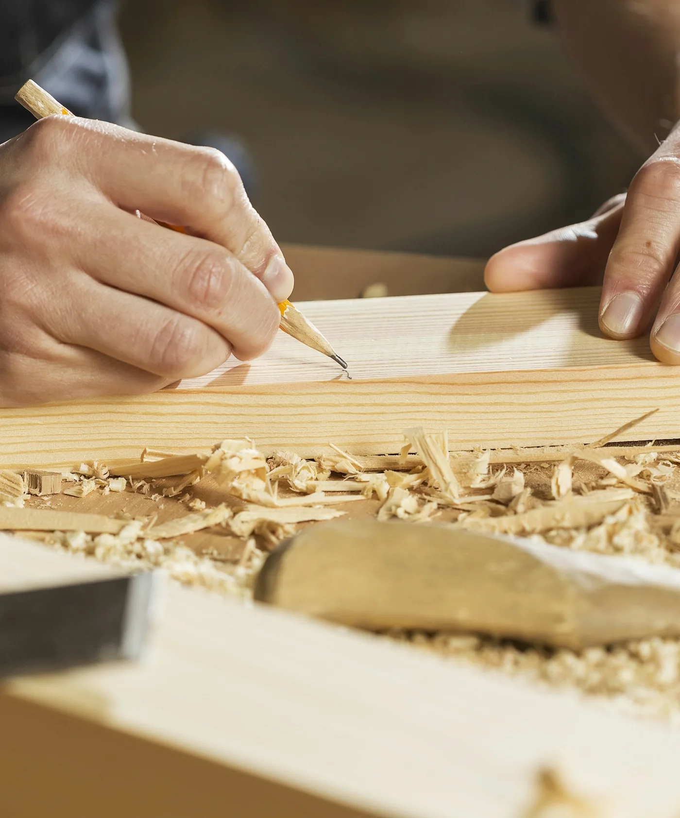 Mains d'artisan traçant un repère au crayon sur une planche, au milieu des copeaux, un ciseau à bois posé devant